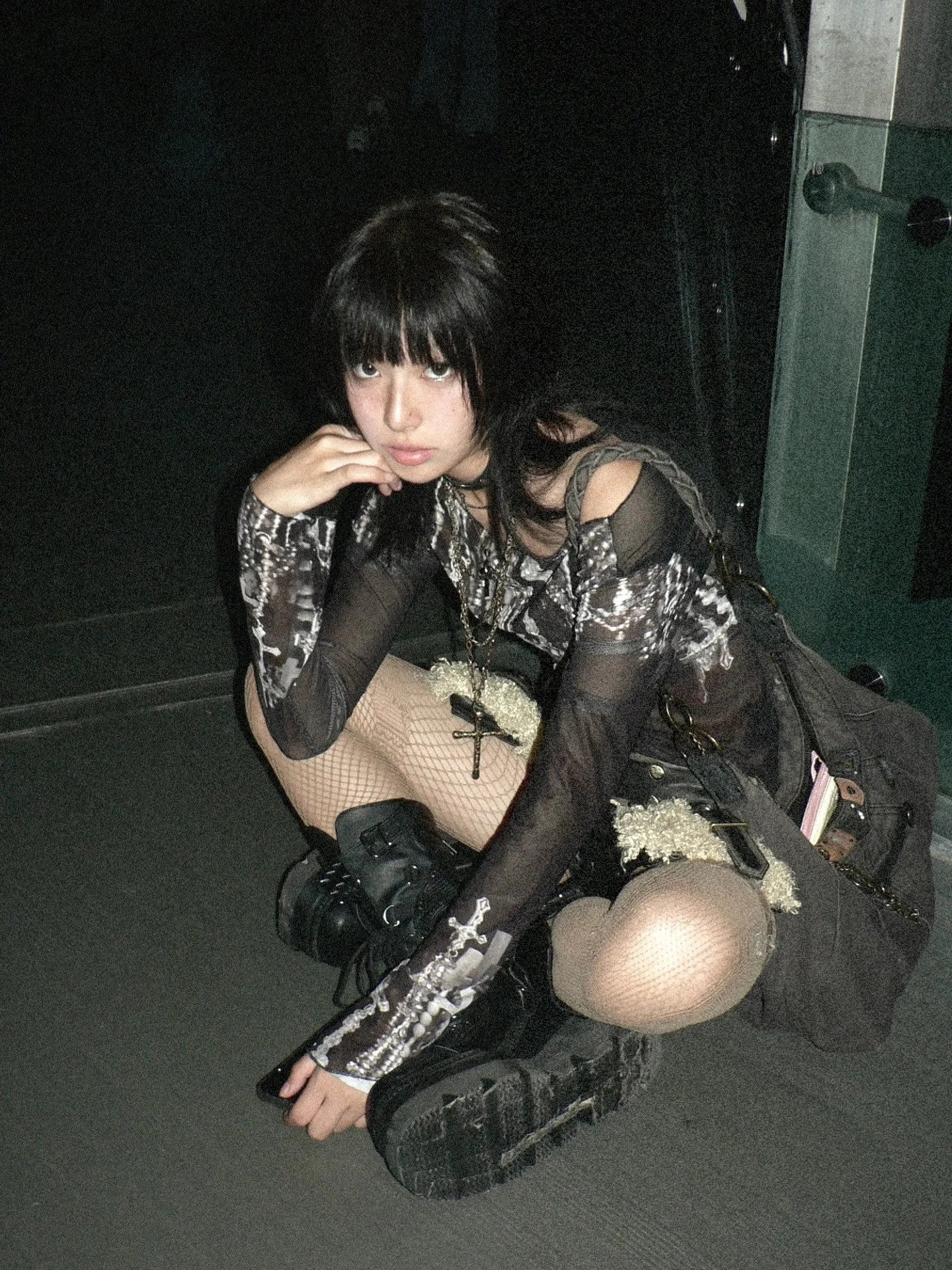 Young woman wearing black affordable emo tops with mesh sleeves and boots crouching indoors